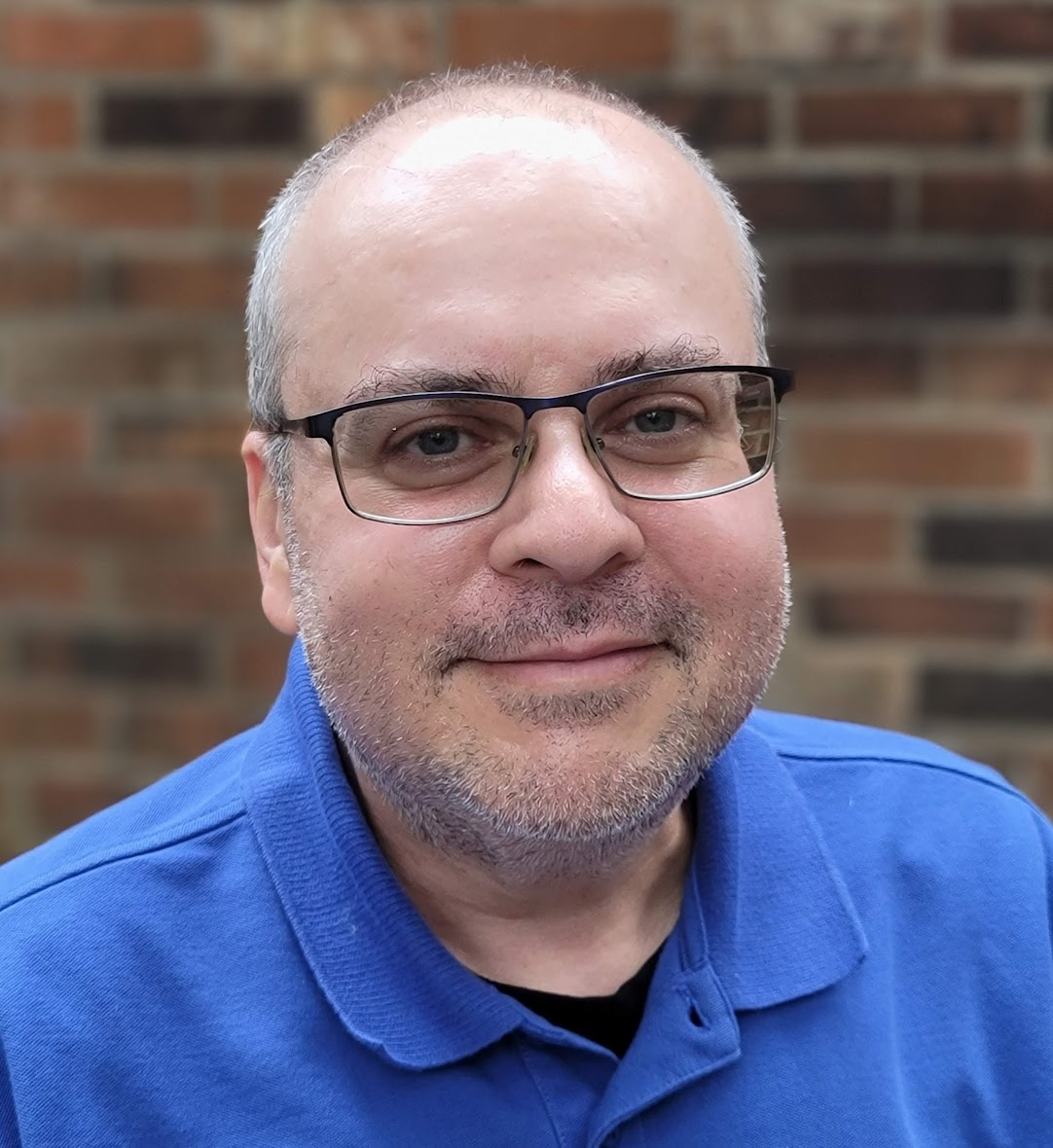 Portrait of Jason A. Feingold wearing glasses and a blue shirt, in front of a brick wall.